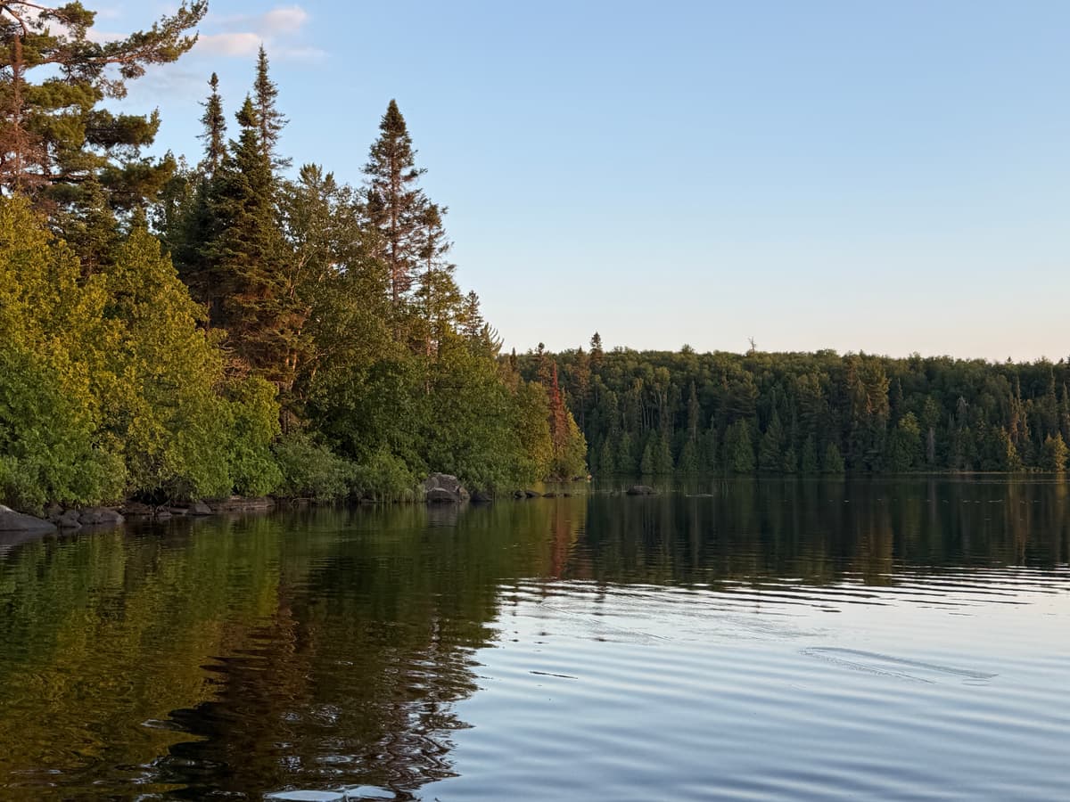 A rocky, tree-lined Boundary Waters shoreline in evening light, the far shore reflected in calm water