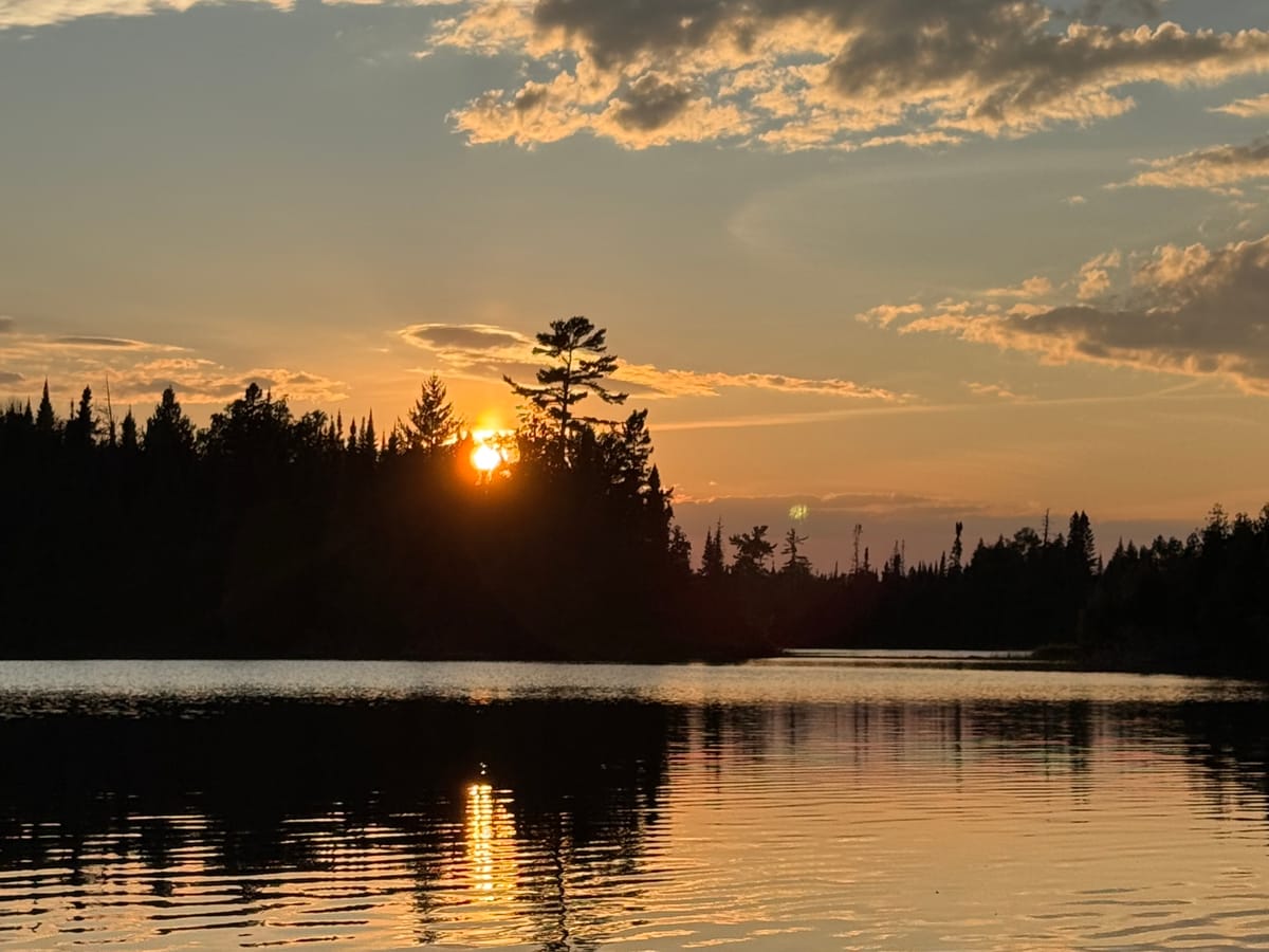 Sunset behind a lone pine on a Boundary Waters shoreline, the sun laying a reflection across the water